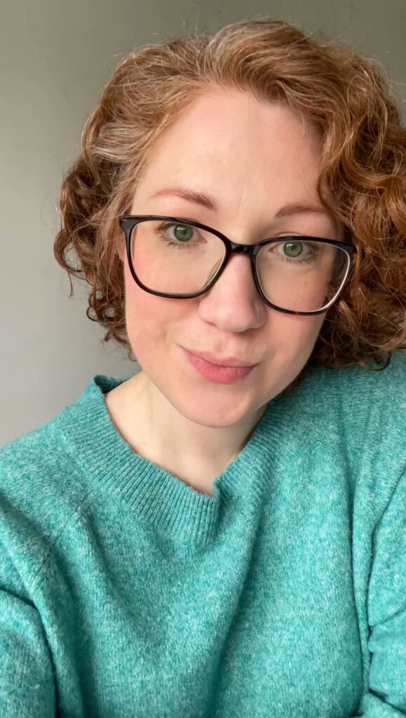 Kerri Awosile Contemporary Feel-good artist from Surrey UK, with her short, auburn, curly hair, glasses, and a turquoise jumper